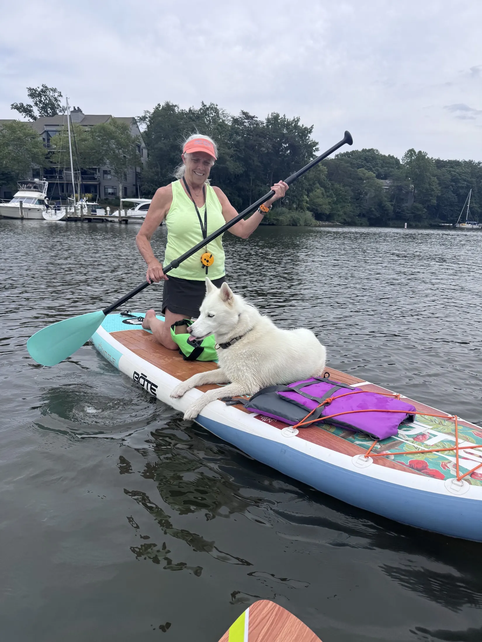 A client kneeling on a paddleboard with her white husky lying calm at the bow, life vest on